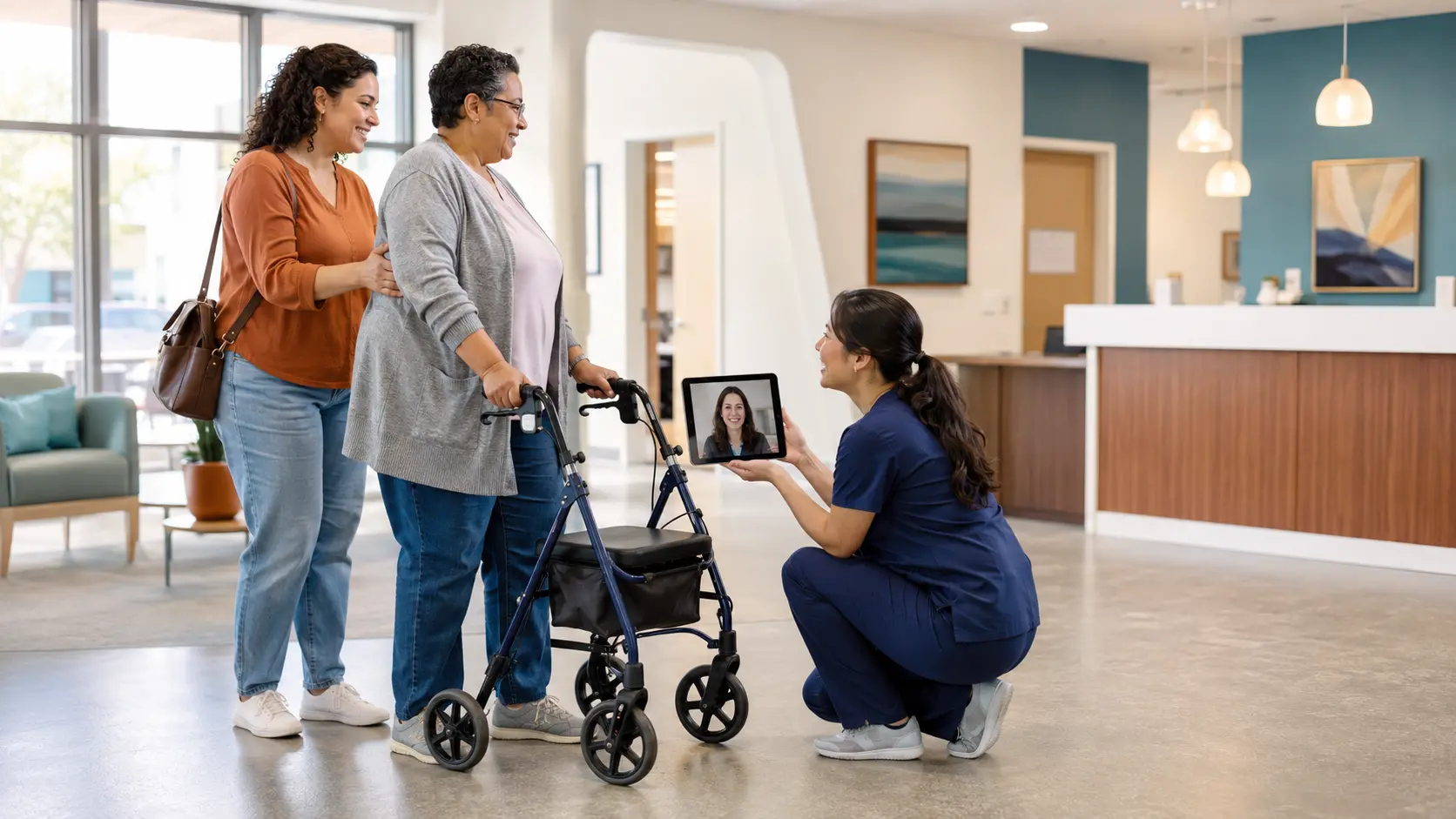 A nurse crouches beside a woman using a rollator and her companion in a hospital lobby while holding a tablet that shows a remote woman on video.