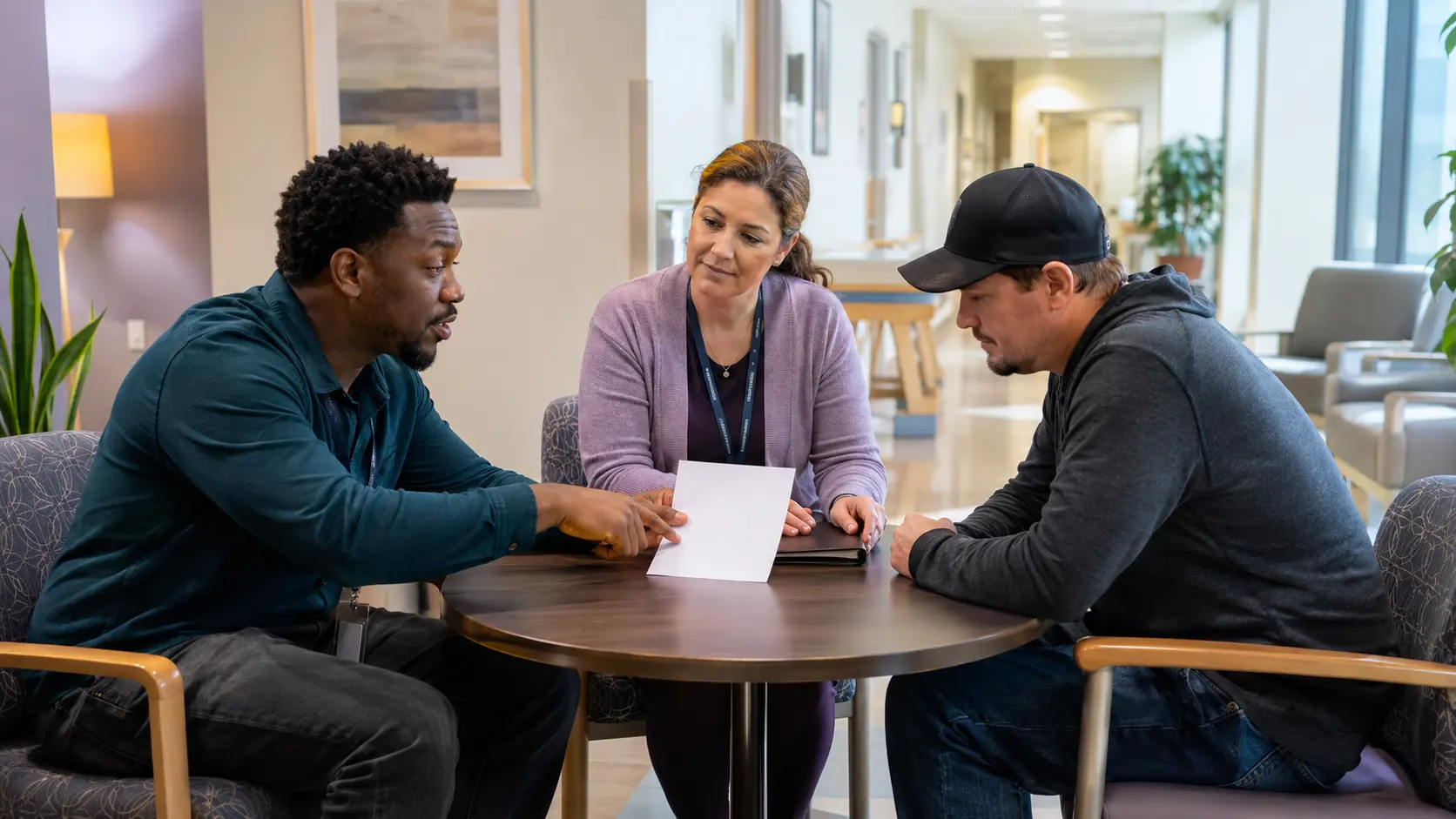 A peer-support navigator and hospital social worker review a blank next-step card with an adult community member in a private transition-of-care area.