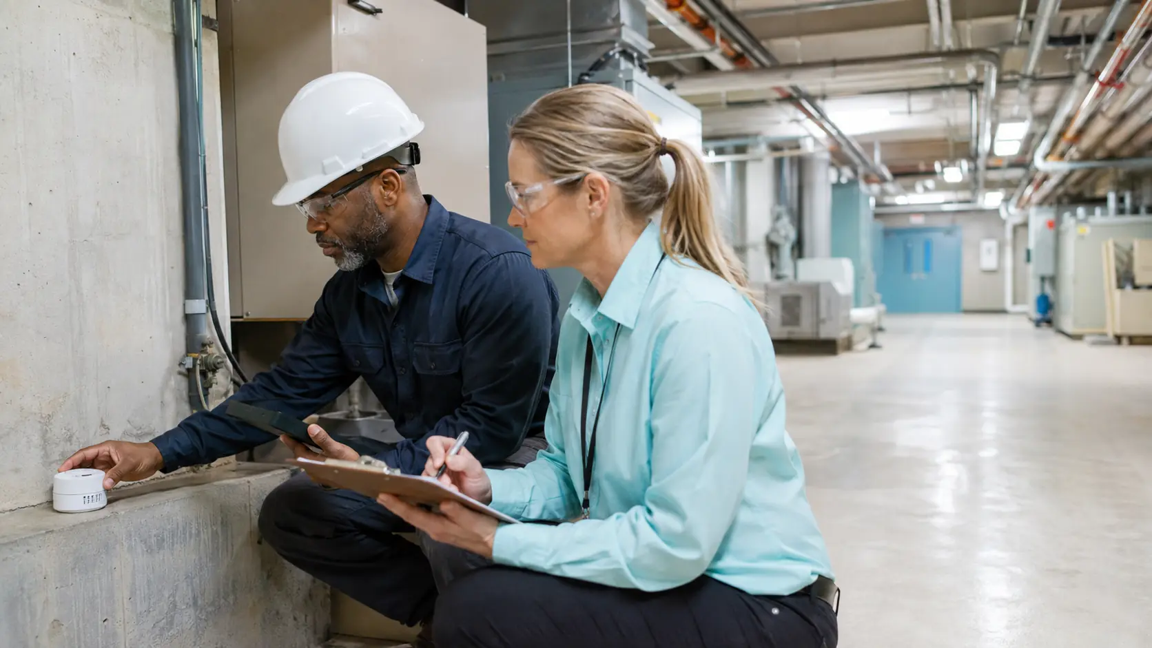 A hospital facilities engineer and environmental health specialist place and document a radon detector in a bright lower-level mechanical space.