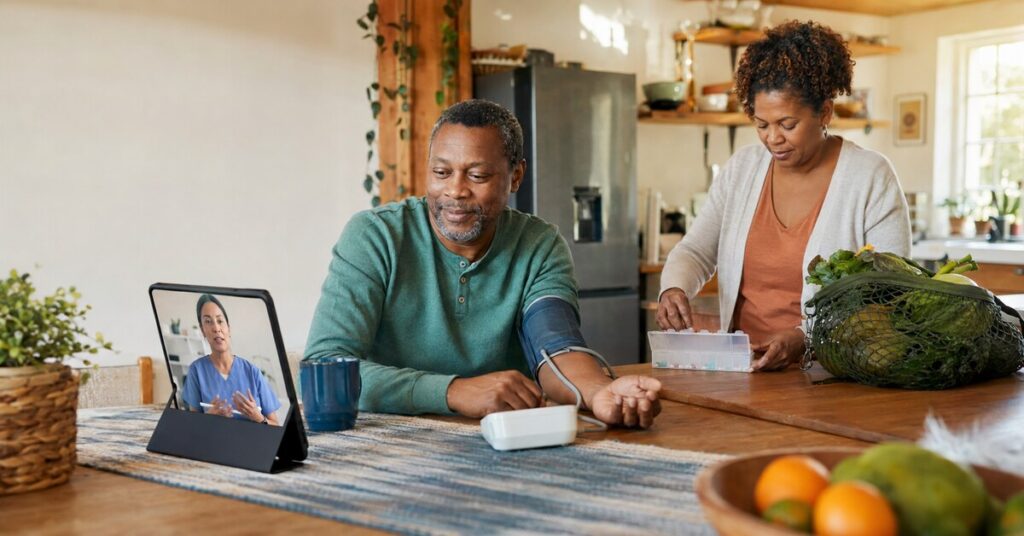 A man checks his blood pressure during a nurse video visit while his partner organizes medication and groceries at home.