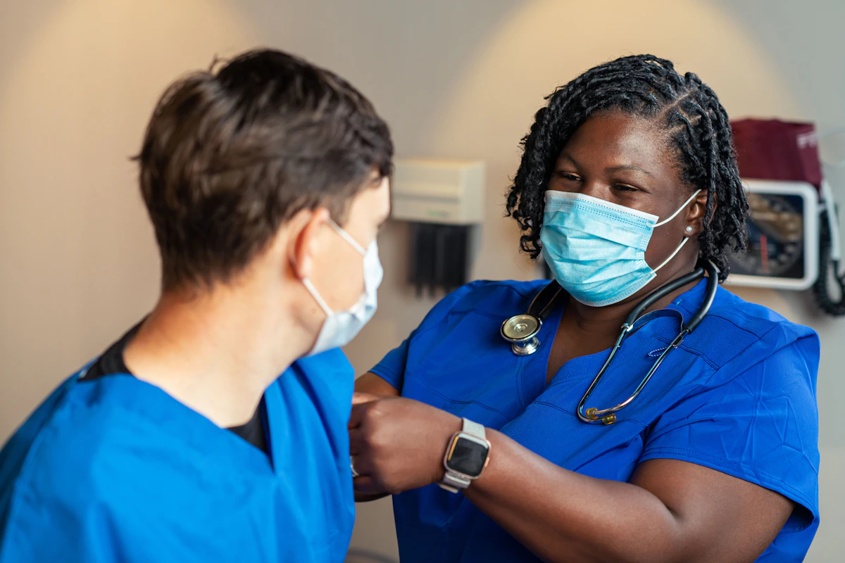 A masked clinician in blue scrubs applies a bandage to a patient's upper arm.